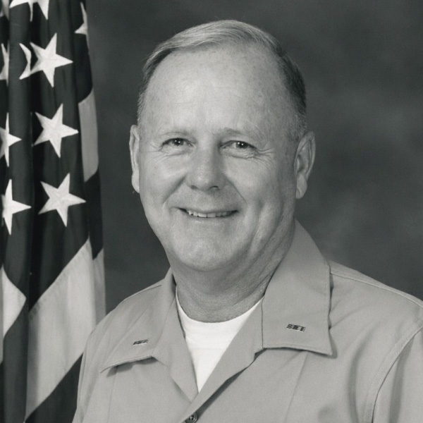 Black and white portrait of Jack O’Neill in military uniform, smiling beside a U.S. flag.